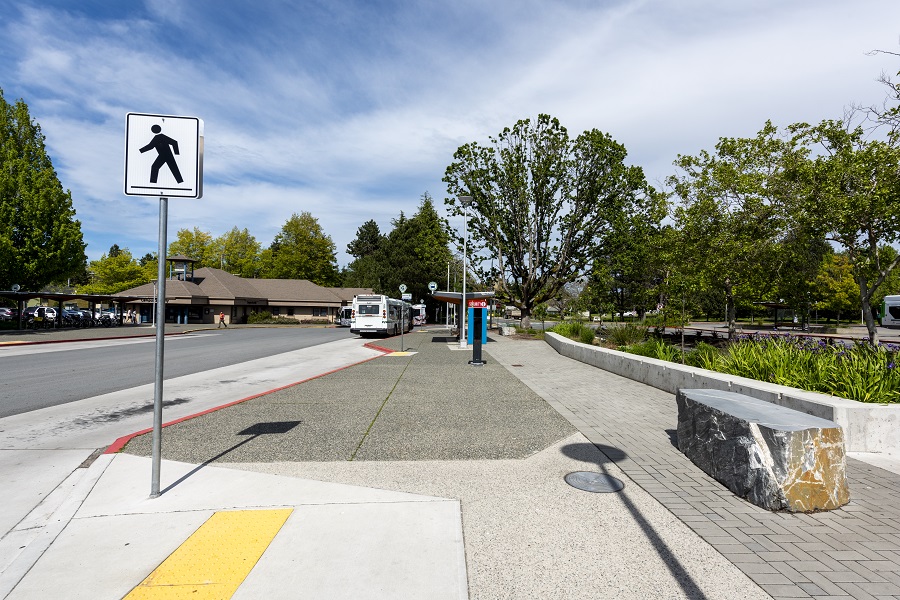 A crosswalk sign is in the foreground. A boulder-style bench is to the side.