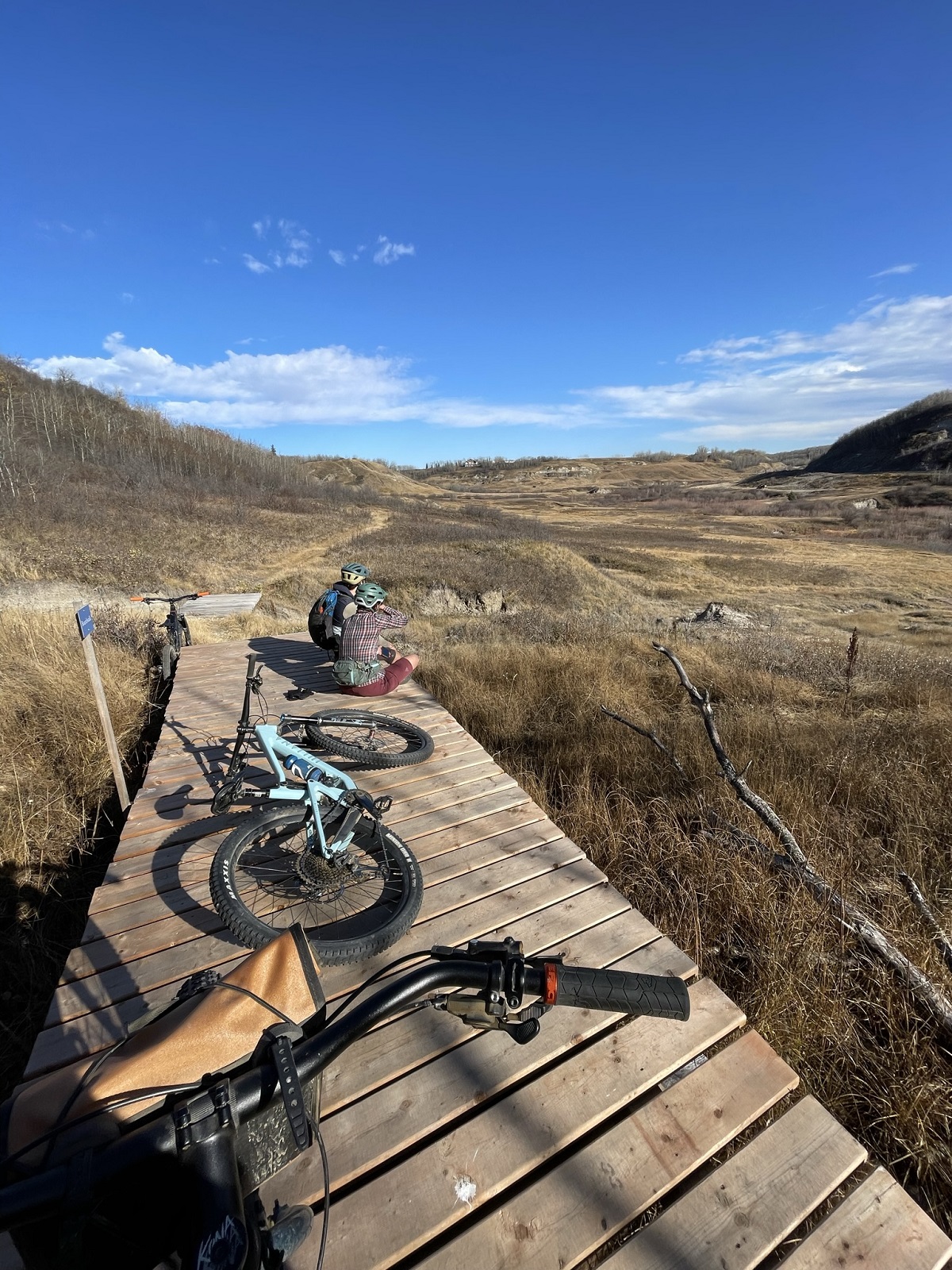 Two mountain bikers are seated on wooden footbridge, their bikes propped and laying around them. The mountain bikers look into the distance. A landscape of rolling grasslands and low-lying wetlands expands in front of them. The sky is a crisp blue.