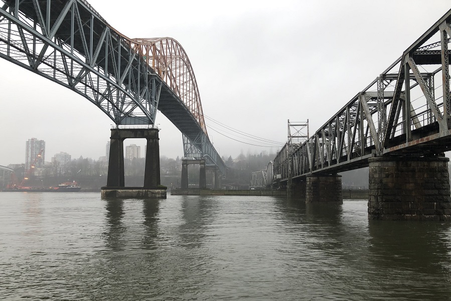 A photo of two bridges, side-by-side, stretching straight ahead into the distance across a river, taken from the perspective of a person on a boat on the water’s surface. The bridges lead to a cityscape that is visible on the far shore of the river. It is cloudy.