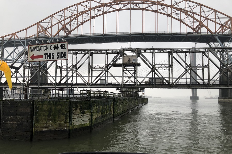 A close-up picture taken of two bridges that span a river, from the perspective of a boat on the river. There is a sign to direct boat traffic under the bridge that reads, “Navigation Channel This Side” with a red arrow pointing to the left. The sky is cloudy and gray. 