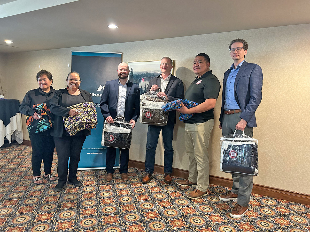 Three Indigenous elders and three McElhanney team members pose for a photograph in a meeting room. All six individuals are holding gifted blankets.