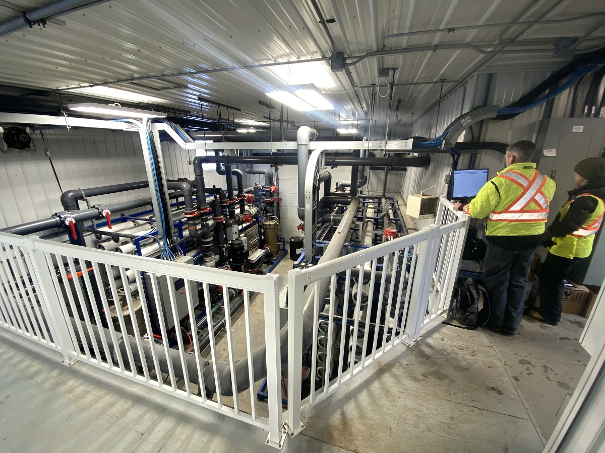 Two individuals in high visibility vests stand at a computer screen on a railed observation deck overlooking the inner workings of a water treatment plant facility.